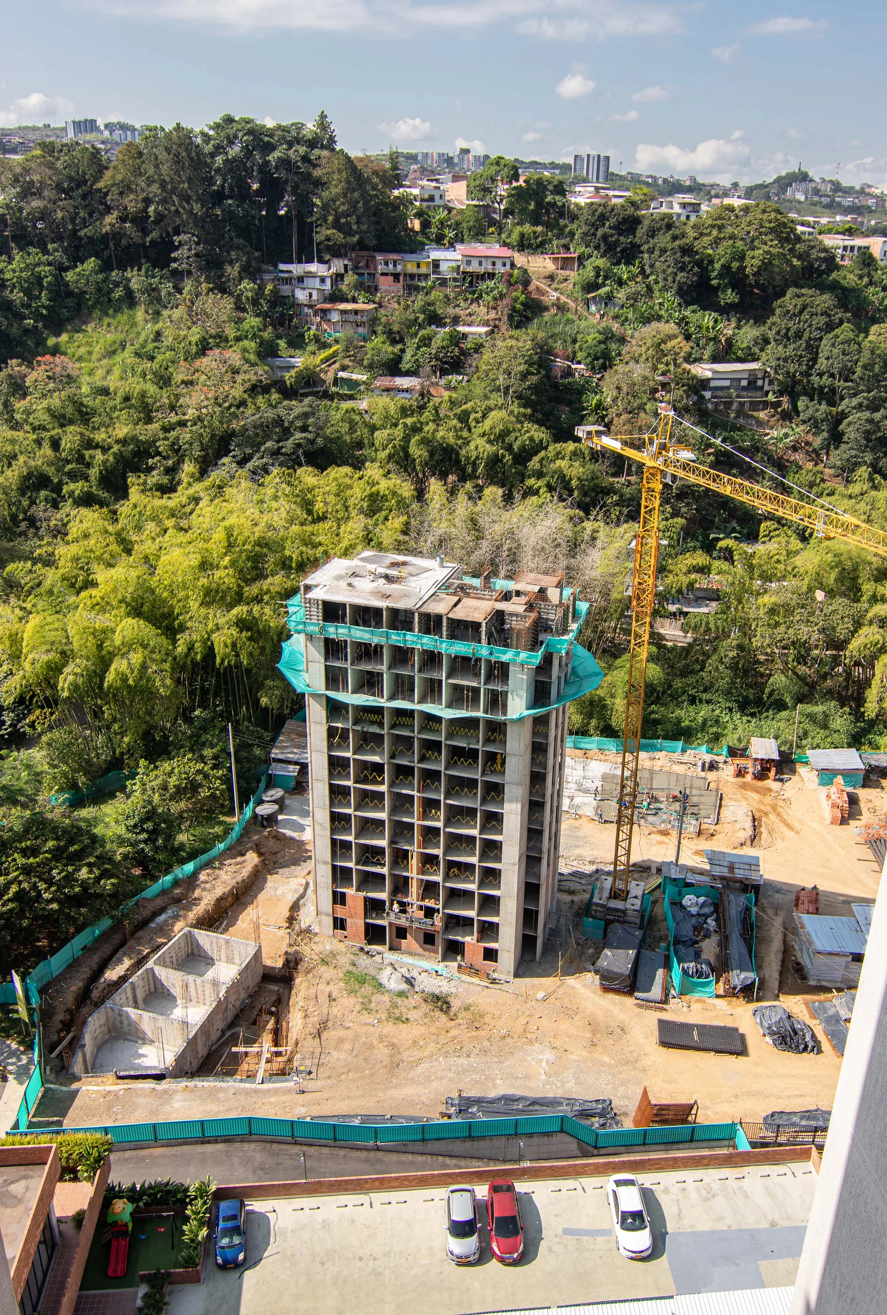 La torre en obra se levanta sobre la ladera de guaduales de Pereira; la grúa y las plataformas escalonadas revelan cómo el proyecto se posa sobre la pendiente.